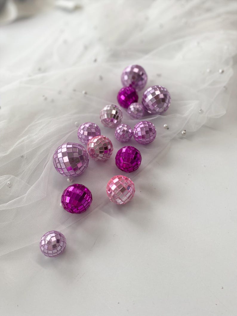Pode incluir: Uma cole&ccedil;&atilde;o de pequenas bolas de discoteca esf&eacute;ricas em v&aacute;rios tons de rosa e roxo, variando do lavanda claro ao magenta profundo. Cada bola &eacute; coberta por min&uacute;sculos espelhos quadrados reflexivos que criam uma superf&iacute;cie facetada.