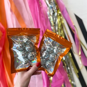 Pode incluir: Dois sacos retangulares de pl&aacute;stico transparente com borda laranja contendo v&aacute;rias pequenas bolas de discoteca prateadas e reflexivas. Cada saco possui um fecho herm&eacute;tico no topo e um acabamento hologr&aacute;fico que reflete a luz.