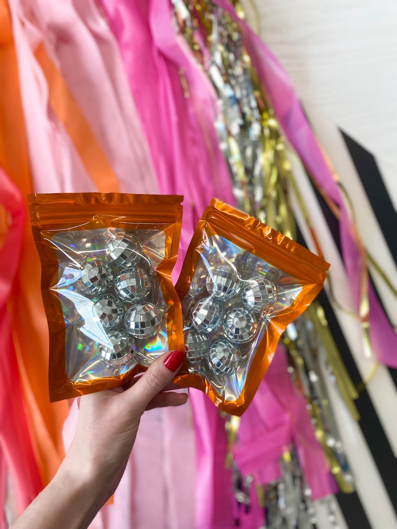 Pode incluir: Dois sacos retangulares de pl&aacute;stico transparente com borda laranja contendo v&aacute;rias pequenas bolas de discoteca prateadas e reflexivas. Cada saco possui um fecho herm&eacute;tico no topo e um acabamento hologr&aacute;fico que reflete a luz.