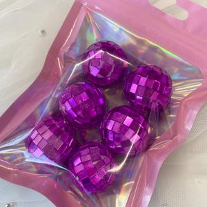 Pode incluir: Um conjunto de seis pequenas bolas de discoteca esf&eacute;ricas com um acabamento refletor roxo vibrante, cada uma coberta com pequenos espelhos quadrados. Estes itens est&atilde;o contidos numa bolsa de pl&aacute;stico transparente e iridescente.