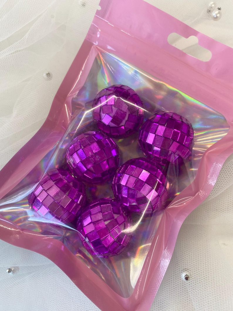 Pode incluir: Um conjunto de seis pequenas bolas de discoteca esf&eacute;ricas com um acabamento refletor roxo vibrante, cada uma coberta com pequenos espelhos quadrados. Estes itens est&atilde;o contidos numa bolsa de pl&aacute;stico transparente e iridescente.