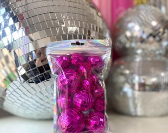 May include: A clear, iridescent resealable plastic bag containing a collection of small, round, hot pink disco balls. Each ball is covered in tiny, reflective square mirrors that catch the light, creating a vibrant and metallic appearance.