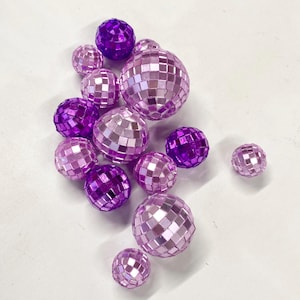 Puede incluir: Una colecci&oacute;n de bolas de discoteca esf&eacute;ricas de varios tama&ntilde;os, con una superficie de mosaico reflectante hecha de peque&ntilde;os espejos cuadrados. Las esferas est&aacute;n coloreadas en tonos morado intenso, lavanda claro y rosa suave.