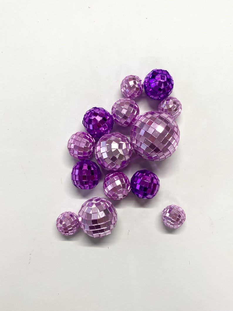 Puede incluir: Una colecci&oacute;n de bolas de discoteca esf&eacute;ricas de varios tama&ntilde;os, con una superficie reflectante compuesta por peque&ntilde;os azulejos de espejo cuadrados de color p&uacute;rpura y rosa claro. El conjunto incluye m&uacute;ltiples esferas de diferentes di&aacute;metros, todas con un acabado met&aacute;lico que capta la luz en sus superficies facetadas.