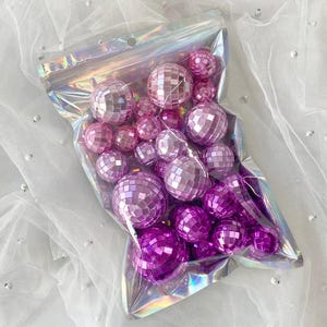 May include: A collection of small, spherical disco balls in varying shades of pink and purple, featuring reflective mirrored facets. These decorative items are contained within a clear, iridescent, resealable plastic pouch with a zip-top closure.