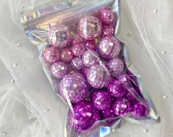 May include: A collection of small, spherical disco balls in varying shades of pink and purple, featuring reflective mirrored facets. These decorative items are contained within a clear, iridescent, resealable plastic pouch with a zip-top closure.