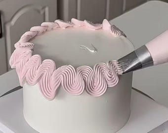 May include: A round white frosted cake being decorated with a piping bag. A metal piping tip is applying a continuous wavy border of light pink buttercream frosting around the top edge of the cake, creating a textured, ruffled design.