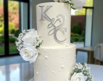 May include: A two-tiered white wedding cake decorated with white roses, baby's breath, and pearl accents. Silver initials "K&C" adorn the top tier. The cake is presented on a white surface.
