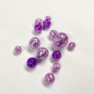 Puede incluir: Una colecci&oacute;n de trece cuentas esf&eacute;ricas tipo bola de discoteca en varios tonos de morado y violeta. Cada cuenta presenta una superficie reflectante y multifacetada compuesta por peque&ntilde;os azulejos de espejo cuadrados y brillantes.