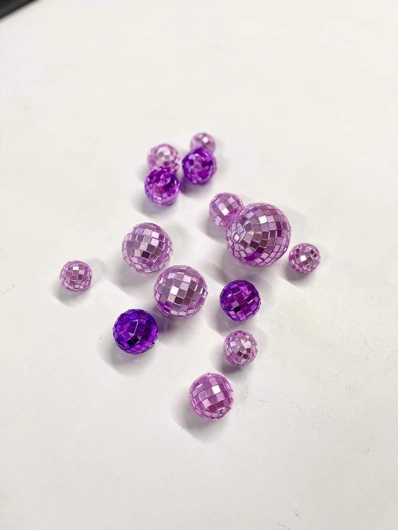Puede incluir: Una colecci&oacute;n de trece cuentas esf&eacute;ricas tipo bola de discoteca en varios tonos de morado y violeta. Cada cuenta presenta una superficie reflectante y multifacetada compuesta por peque&ntilde;os azulejos de espejo cuadrados y brillantes.