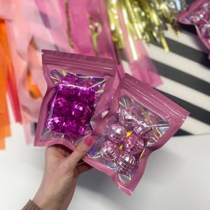Puede incluir: Dos peque&ntilde;as bolsas rosas resellables que contienen m&uacute;ltiples adornos de bolas de discoteca facetadas y reflectantes. Una bolsa contiene bolas de color magenta intenso, mientras que la otra contiene bolas de color rosa claro, ambas con una superficie brillante.