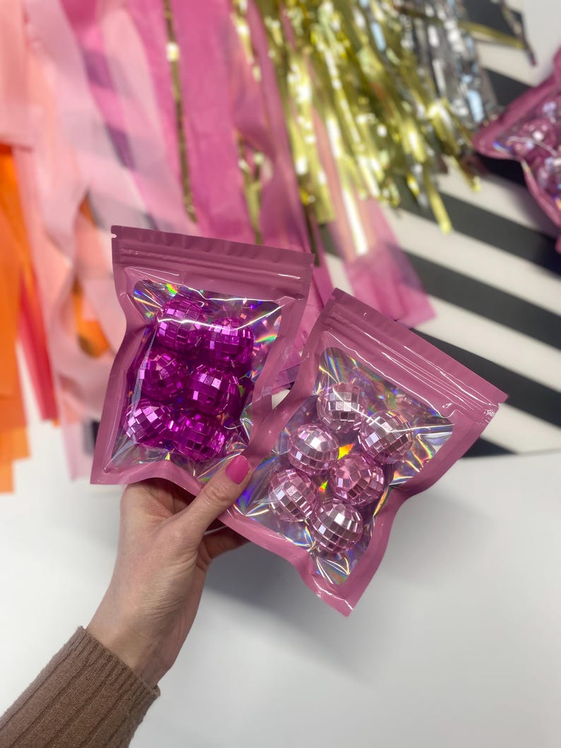 Puede incluir: Dos peque&ntilde;as bolsas rosas resellables que contienen m&uacute;ltiples adornos de bolas de discoteca facetadas y reflectantes. Una bolsa contiene bolas de color magenta intenso, mientras que la otra contiene bolas de color rosa claro, ambas con una superficie brillante.
