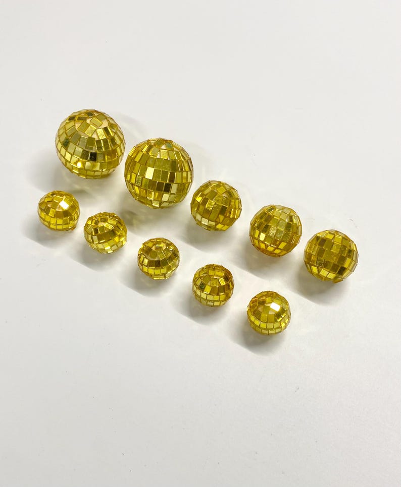 Puede incluir: Un conjunto de diez bolas de discoteca esf&eacute;ricas de distintos tama&ntilde;os, cubiertas con peque&ntilde;os espejos cuadrados dorados y reflectantes. Las esferas est&aacute;n dispuestas en un grupo suelto, mostrando el acabado dorado met&aacute;lico y el patr&oacute;n de cuadr&iacute;cula.