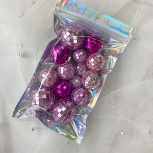 Pode incluir: Uma cole&ccedil;&atilde;o de pequenas bolas de discoteca esf&eacute;ricas em v&aacute;rios tons de rosa e roxo, contidas numa bolsa de pl&aacute;stico transparente, iridescente e sel&aacute;vel. As bolas apresentam uma superf&iacute;cie refletora composta por pequenos espelhos.