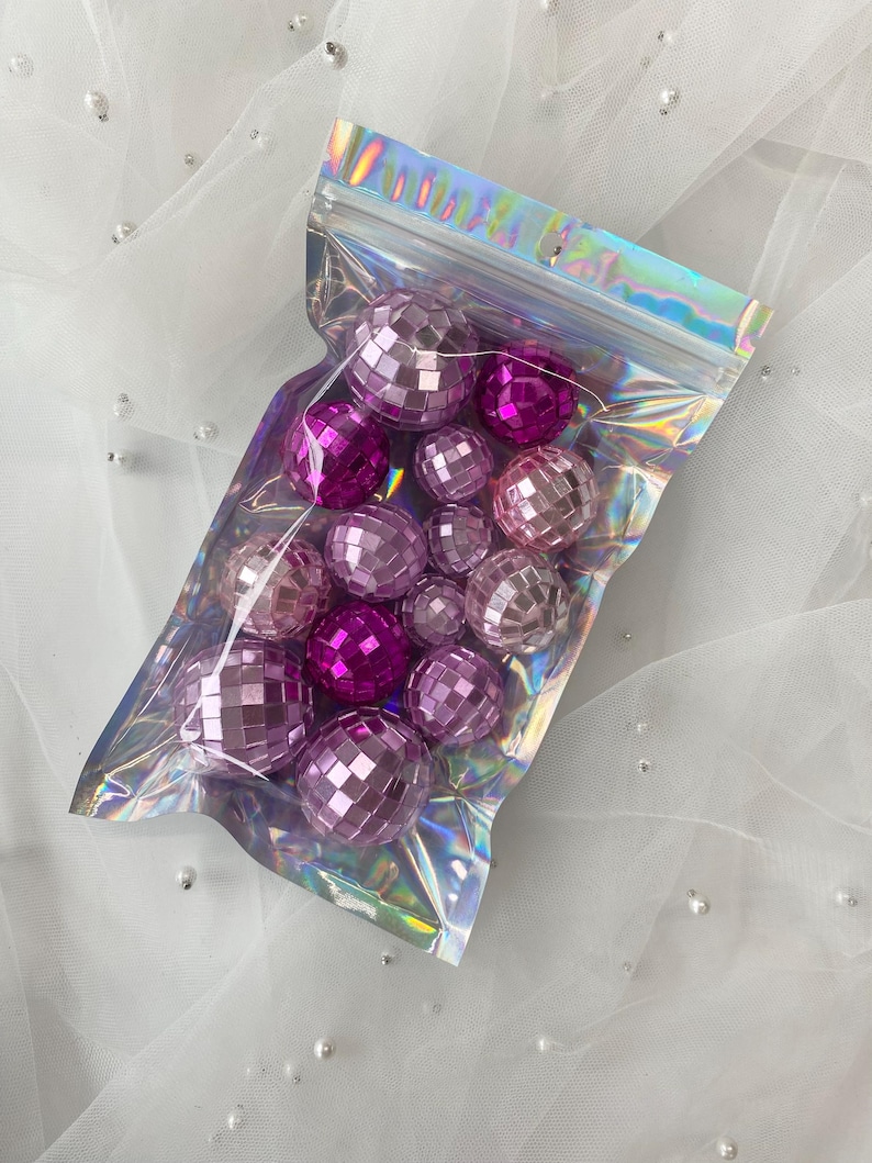 Pode incluir: Uma cole&ccedil;&atilde;o de pequenas bolas de discoteca esf&eacute;ricas em v&aacute;rios tons de rosa e roxo, contidas numa bolsa de pl&aacute;stico transparente, iridescente e sel&aacute;vel. As bolas apresentam uma superf&iacute;cie refletora composta por pequenos espelhos.