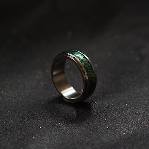 May include: A silver ring with a green band featuring a swirling pattern. The ring is on a black surface.