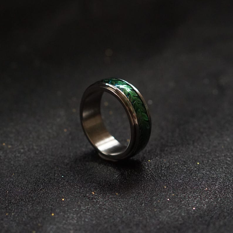May include: A silver ring with a green band featuring a swirling pattern. The ring is on a black surface.