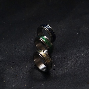 May include: Three stainless steel rings with textured bands in blue, green, and gold. The rings are stacked diagonally, showcasing their metallic finish and colorful accents. The rings are likely fashion accessories.