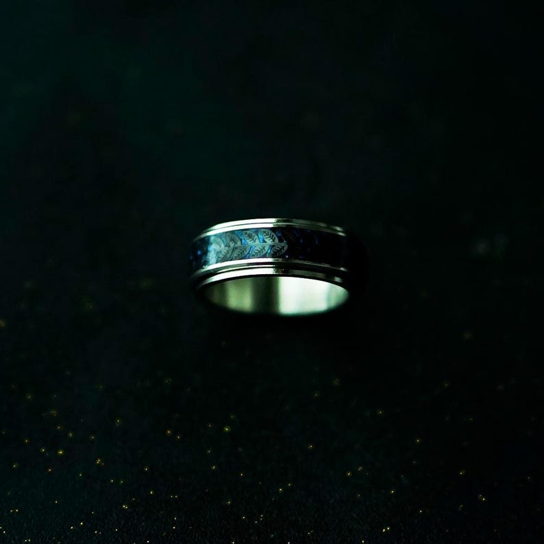 May include: A silver ring with a dark blue band featuring a swirling, iridescent pattern of blues and greens. The ring has a polished, metallic finish and a wide band design. The ring is set against a dark background.
