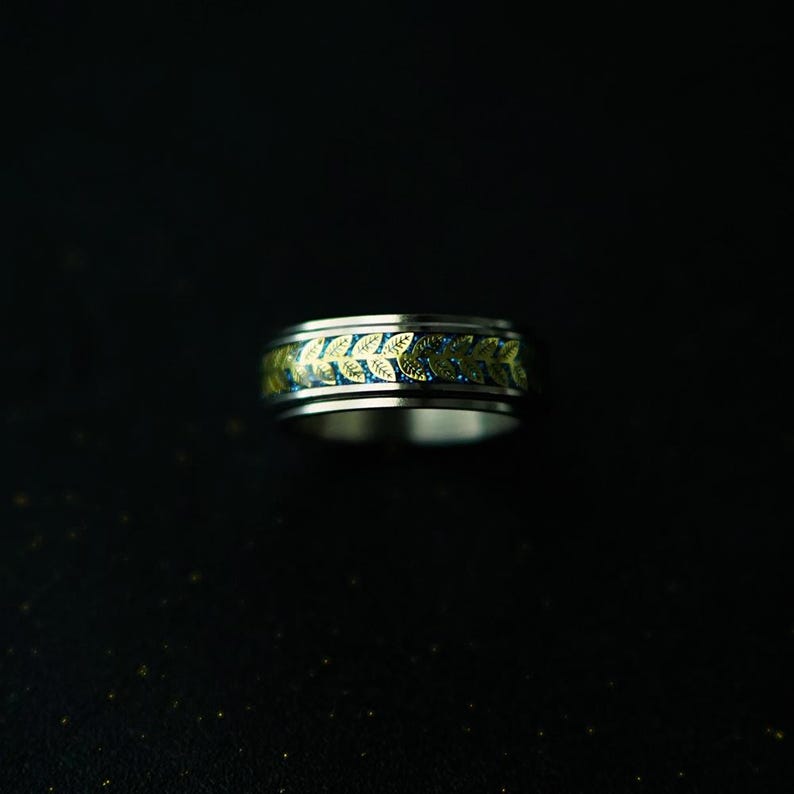 May include: A silver ring with a gold leaf pattern encircling a blue band. The ring has a polished finish and a detailed design, suitable for everyday wear or special occasions.
