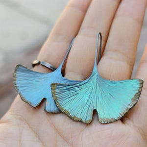 May include: A pair of teal-colored leaf-shaped earrings. The earrings have a textured surface with a metallic sheen and a gold-colored edge. The earrings are held in a hand.
