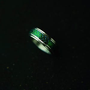 May include: A silver ring with a green leaf pattern. The ring features a band of green leaves set against a blue background. The ring is a piece of jewelry.