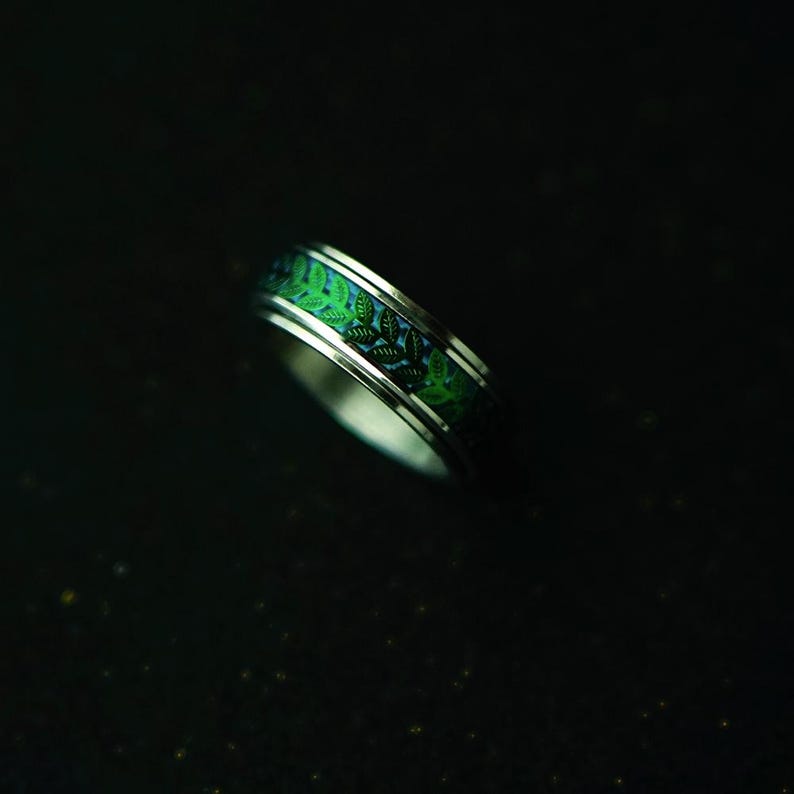May include: A silver ring with a green leaf pattern. The ring features a band of green leaves set against a blue background. The ring is a piece of jewelry.