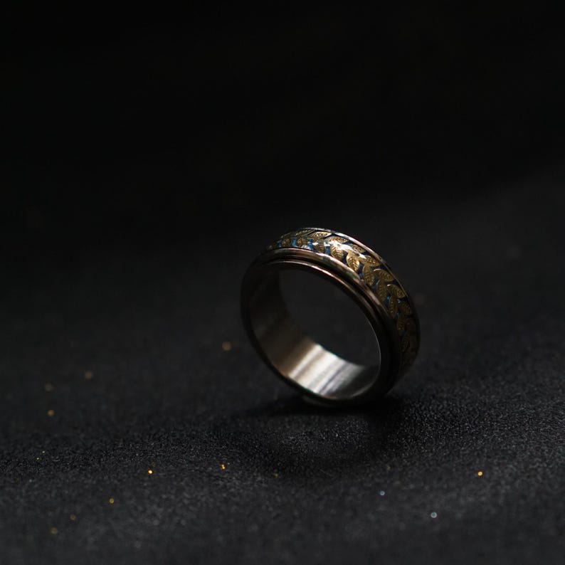 May include: A silver ring with a gold and blue patterned band. The ring is on a black surface.