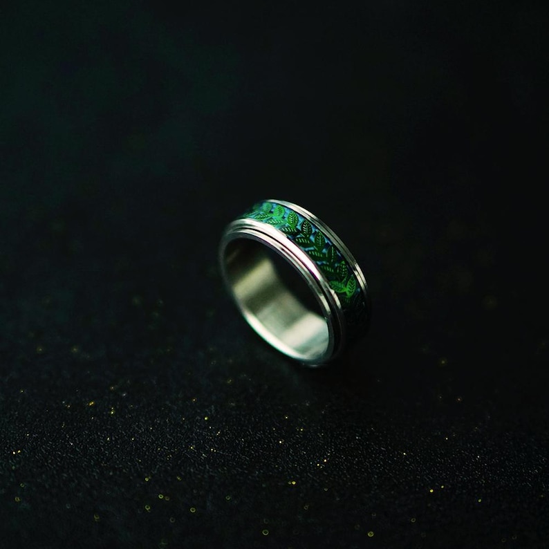 May include: A silver ring with a green and blue patterned band. The ring has a polished finish and a detailed design. The band features a repeating leaf pattern in shades of green and blue. The ring is set against a dark background.