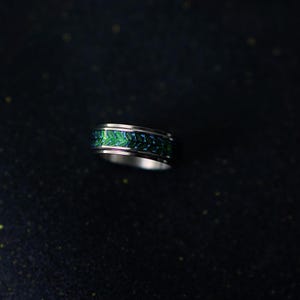 May include: A silver ring with a green and blue patterned band. The band features a repeating design of leaves or feathers. The ring is set against a dark background.