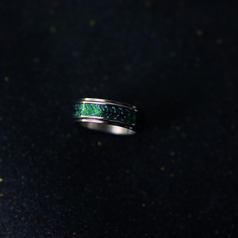 May include: A silver ring with a green and blue patterned band. The band features a repeating design of leaves or feathers. The ring is set against a dark background.