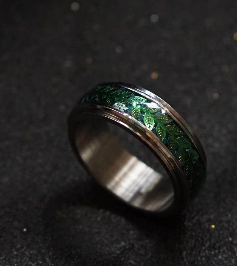May include: A silver-toned ring with a band featuring a green leaf design. The ring has a polished finish and a detailed pattern of leaves. The ring is a piece of jewelry.