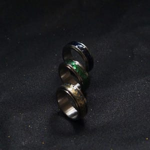 May include: Three silver spinning rings with different colored inlays. The rings are stacked on top of each other. The top ring has a dark blue inlay, the middle ring has a green inlay, and the bottom ring has a gold inlay.