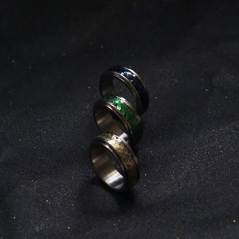 May include: Three silver spinning rings with different colored inlays. The rings are stacked on top of each other. The top ring has a dark blue inlay, the middle ring has a green inlay, and the bottom ring has a gold inlay.