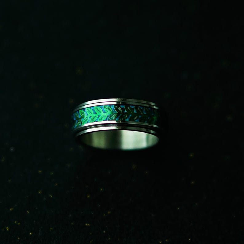 May include: A silver ring with a band featuring a repeating pattern of green leaves on a blue background. The ring has a polished, metallic finish and is set against a dark background.