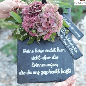 Könnte beinhalten: Ein Blumenarrangement mit rosa und lila Hortensien und anderen Blumen auf einer dunklen Schiefertafel. Die Tafel trägt den weißen Text: „Deine Reise endet hier, aber nicht die Erinnerungen, die du uns geschenkt hast!“ und „In stiller, liebevoller Erinnerung“.