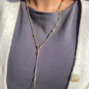 May include: A gold-coloured necklace with a Y-shaped pendant. The necklace features small, colourful beads in red, blue, yellow, and green. The necklace is layered with a second, thinner gold chain. The person is wearing a grey shirt and a white cardigan.