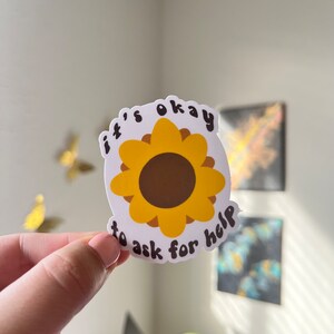 May include: A white sticker with a yellow sunflower design and the text "it's okay to ask for help" in black. The sunflower has a brown center and yellow petals. The sticker is held up against a white background.