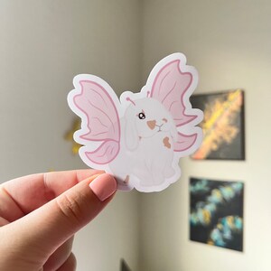 May include: A white rabbit with pink butterfly wings. The rabbit has brown spots and is looking to the right. The wings are a light pink and have a delicate, whimsical feel.