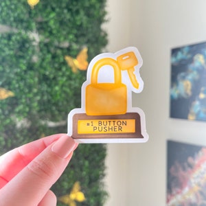 May include: A gold-colored padlock with a key in the keyhole, sitting on a brown base with the text "#1 BUTTON PUSHER".