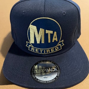 May include: A navy blue baseball cap with a gold embroidered MTA logo and the word "Retired" underneath. The cap has a black and silver "Snapback" label on the back.