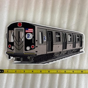 Puede incluir: Un parche bordado de color gris y negro que representa un tren de metro de la ciudad de Nueva York con el logotipo de la MTA y el número 2 en el lateral.