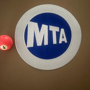 May include: A circular patch featuring the letters "MTA" in white against a blue background, encircled by a white border. A red and orange billiard ball with the number "4" is next to the patch. The patch is on a brown surface.