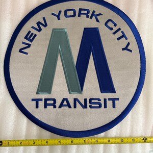 以下が含まれることがあります： 青色の「NEW YORK CITY」の文字が円形に配置され、中央のデザインを囲んでいます。デザインは、青と緑の色合いの大きな「M」と、その下に「TRANSIT」の文字が特徴です。パッチの直径は約30cmです。