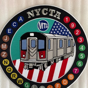 May include: A round, black and white patch with the text "NYCTA" in silver lettering. The patch features a stylized image of a subway train with the text "MTA" in a blue circle. The train is on a red and white striped background. The numbers 1 through 7 and the letters A through W are arranged in a circle around the train.