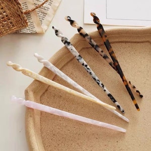 May include: Five hair sticks with a twisted design. The sticks are made of a marbled plastic in various colors, including black, white, brown, and pink.