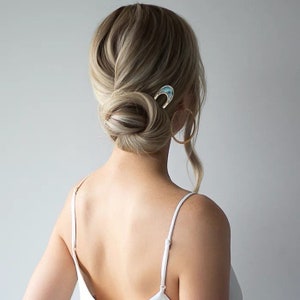 May include: A woman with blonde hair styled in a low bun with a gold and blue hair clip. She is wearing a white tank top and large gold hoop earrings.