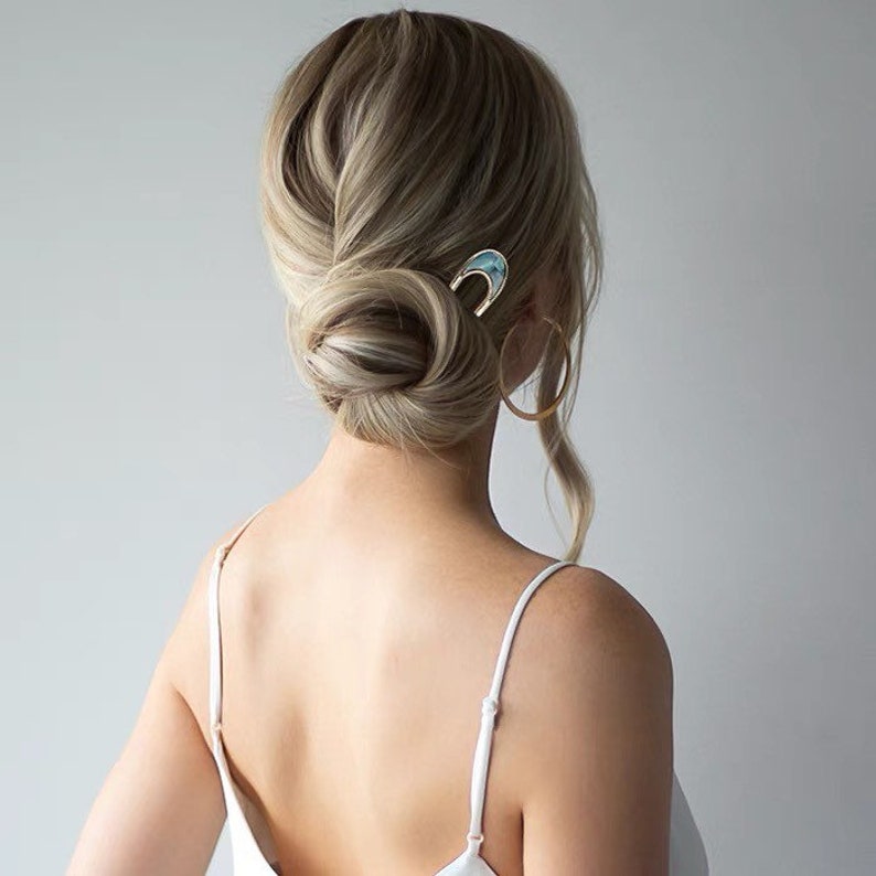 May include: A woman with blonde hair styled in a low bun with a gold and blue hair clip. She is wearing a white tank top and large gold hoop earrings.