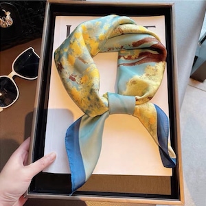 May include: A square silk scarf with a watercolor design in shades of blue, yellow, and brown. The scarf is tied in a knot, with the ends hanging down. The scarf is displayed in a black box.