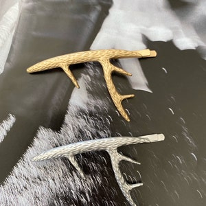 Peut inclure: Deux pinces &agrave; cheveux en forme de bois de cerf. L'une est dor&eacute;e, l'autre argent&eacute;e. Les deux pinces ont une surface textur&eacute;e et une fermeture de type barrette. Les pinces sont pr&eacute;sent&eacute;es sur un fond noir et blanc.